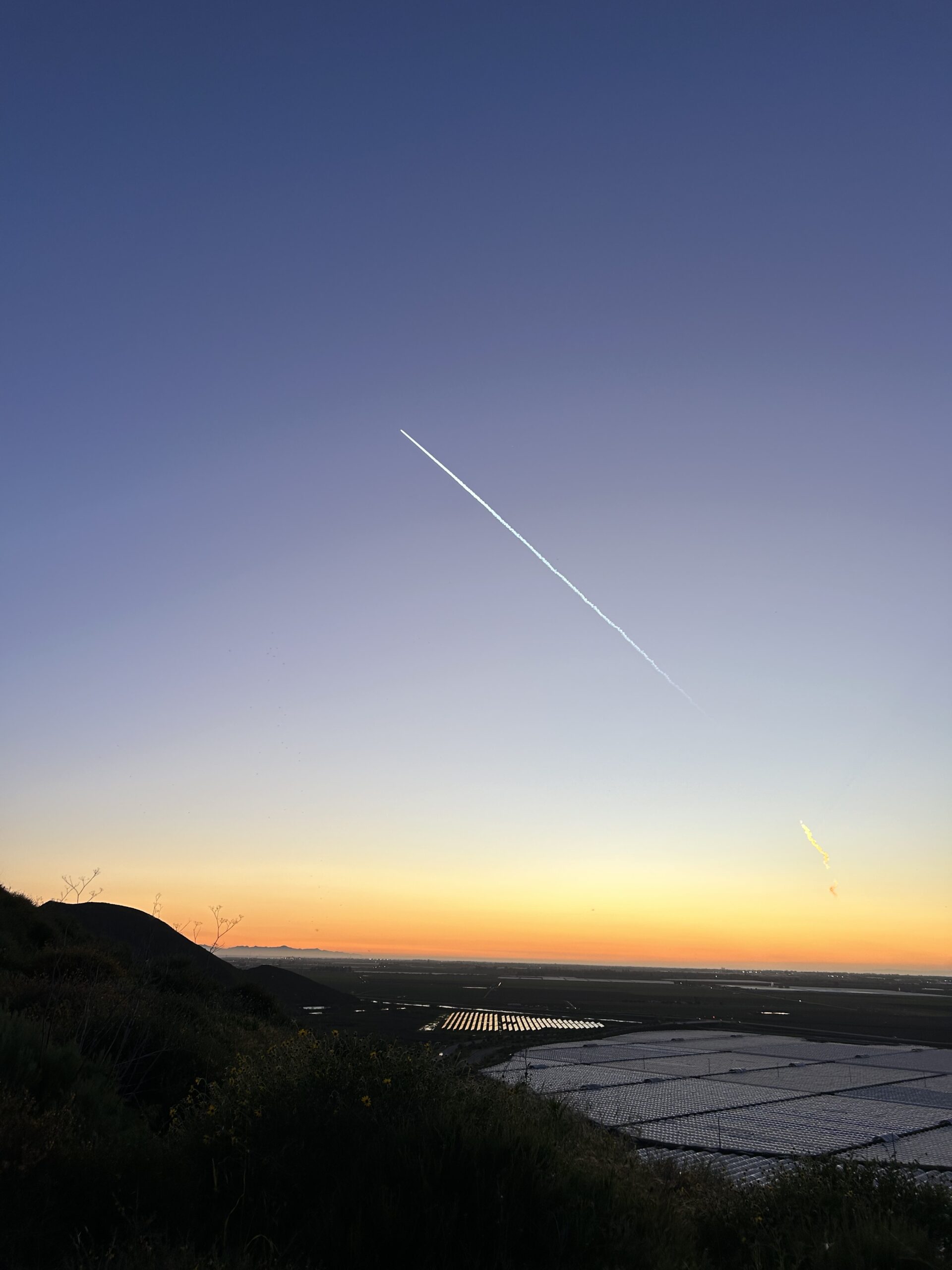 Sunset view over farms and the pacific ocean. A large streak crosses the twilight sky. At the upper edge of the streak is a SpaceX rocket.